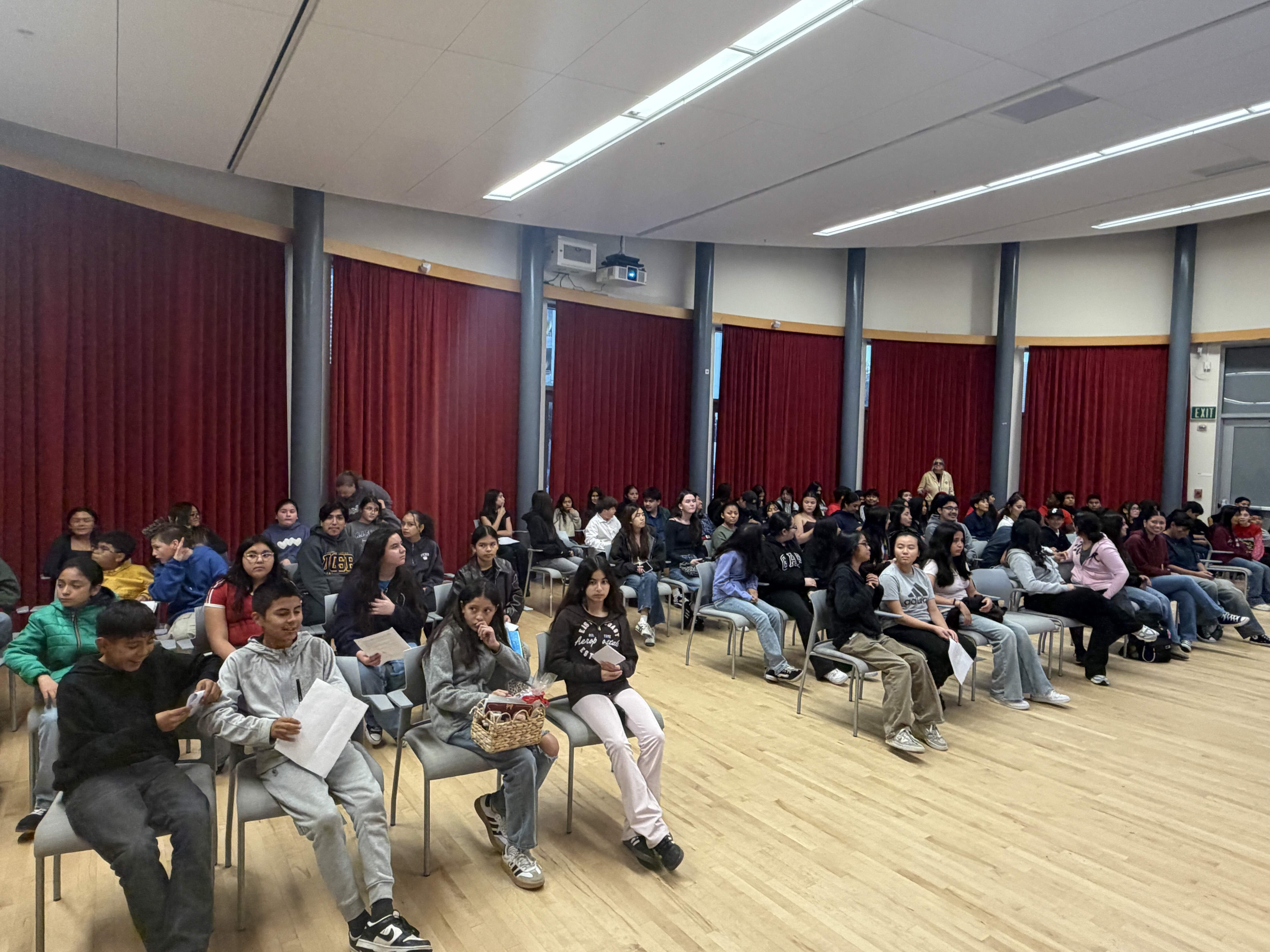 A wide shot of a lecture hall showing a large group of diverse students seated in grey chairs, facing forward attentively. The room is circular with floor-to-ceiling red curtains along the walls. Some students in the front row are holding papers or small baskets.