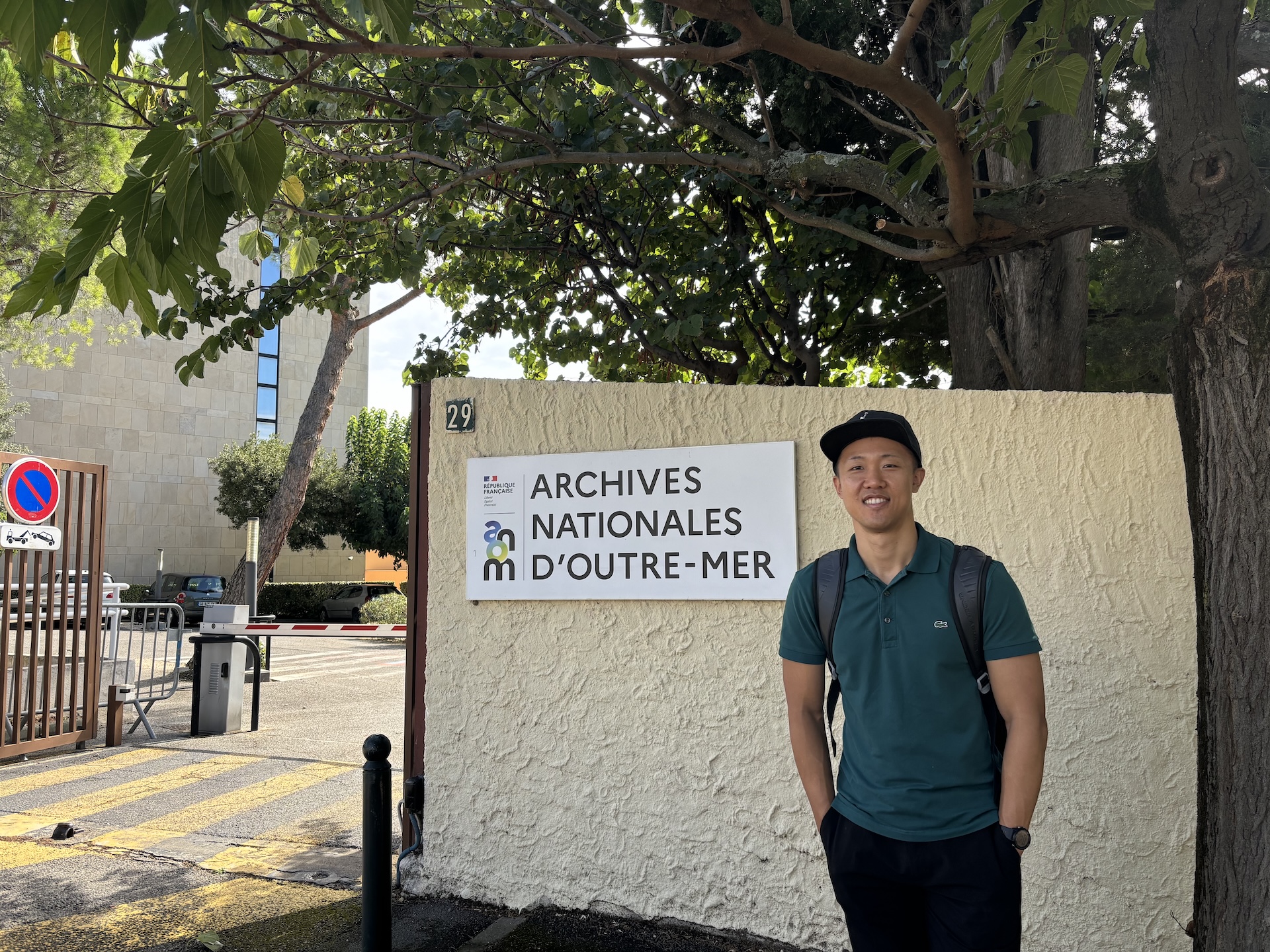 Doctoral candidate Jungki Min smiling and standing near the outdoor entrance sign for the Archives Nationales d'Outre-Mer in France.