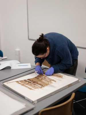 Doctoral candidate Alida Jekabson, wearing blue nitrile gloves, carefully examines and measures a historical Andean khipu, a knotted-fiber record-keeping device, laid flat on a table in a research lab.