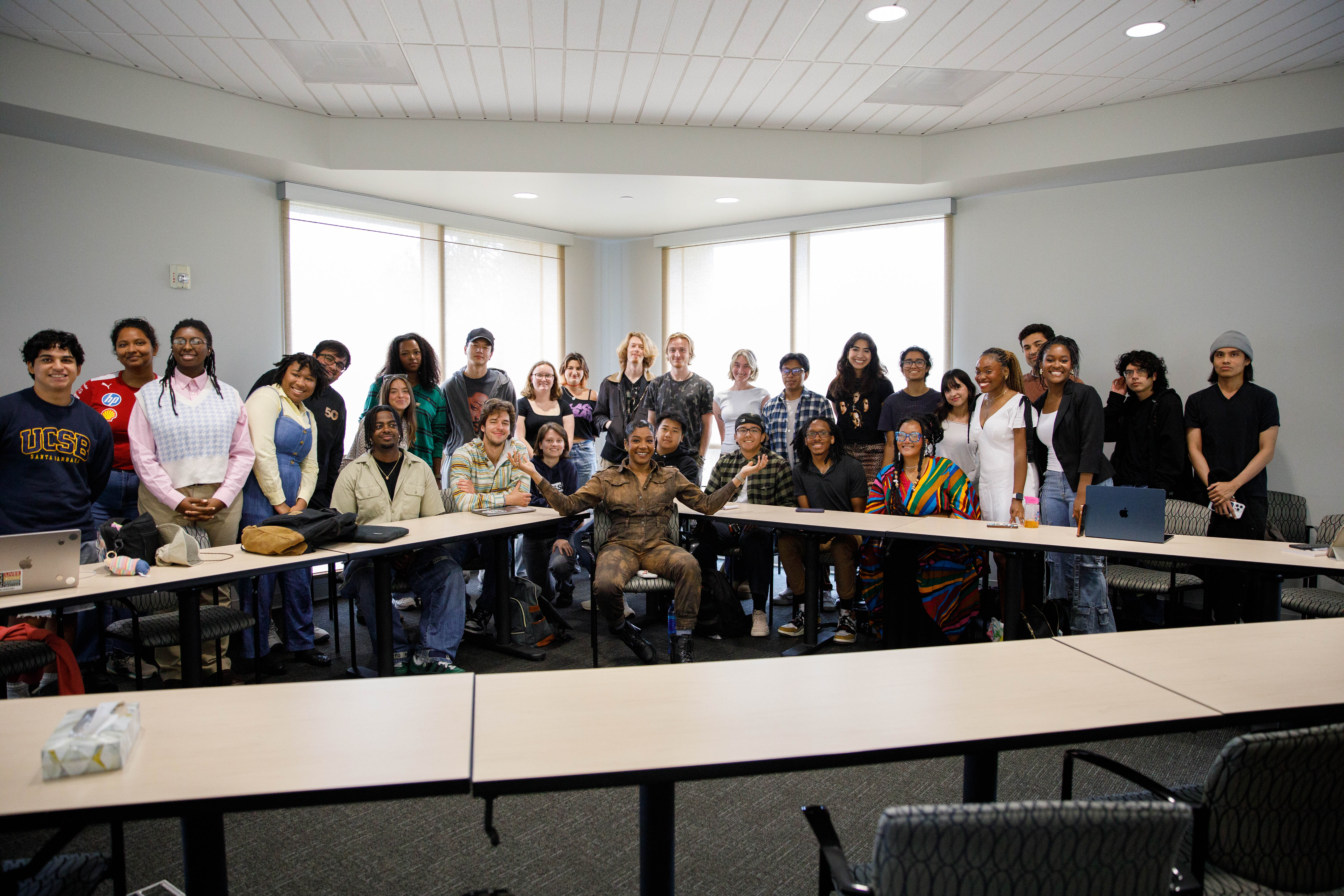 A joyful group photograph of comedian Tiffany Haddish with dozens of diverse university students in a conference room. Haddish is seated in the center, wearing a khaki jumpsuit and smiling, while the students are gathered closely behind and around a U-shaped arrangement of tables.