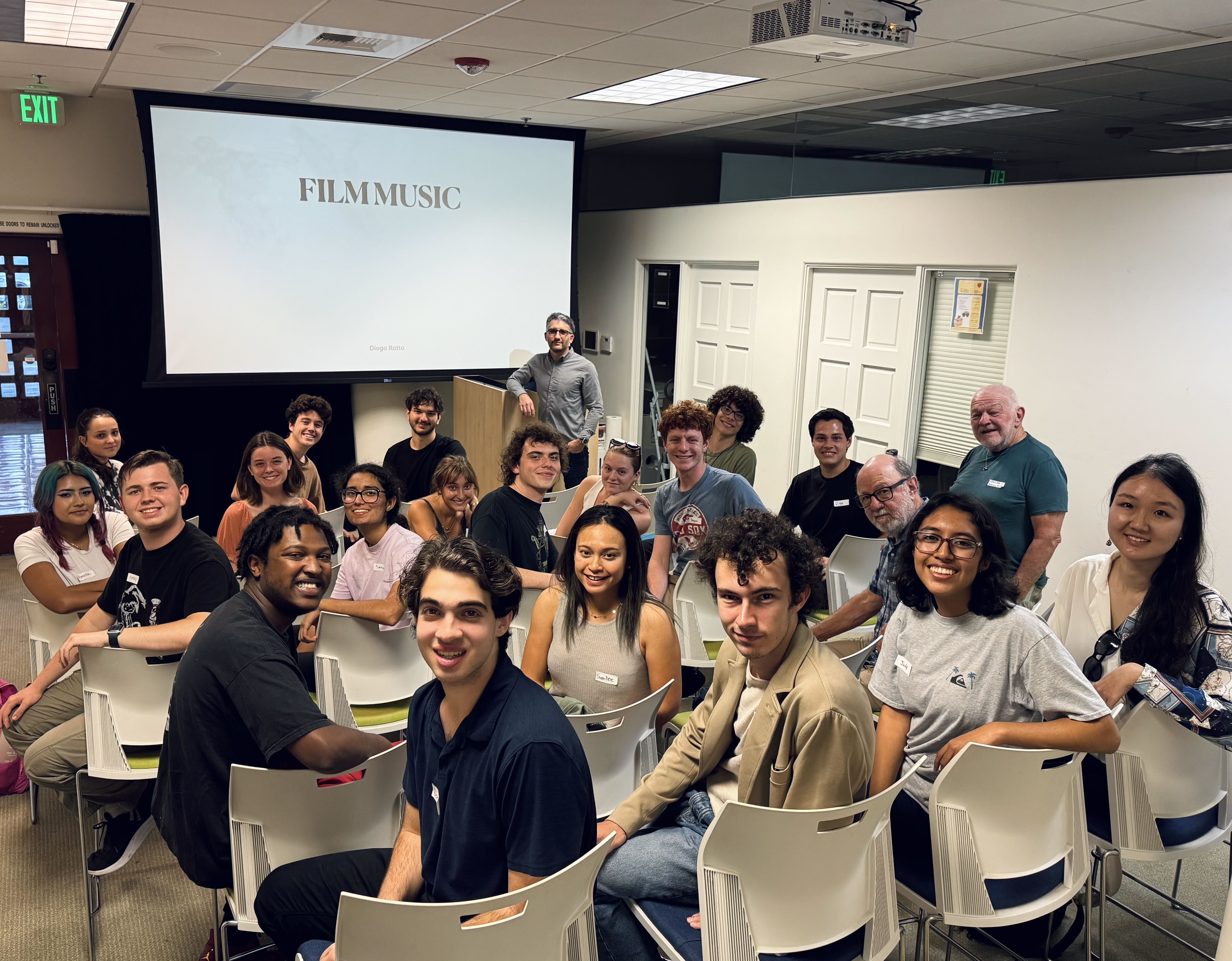 A large group of UCSB students and instructors smile for a photo inside a classroom with a projector screen reading "FILM MUSIC" in the background.