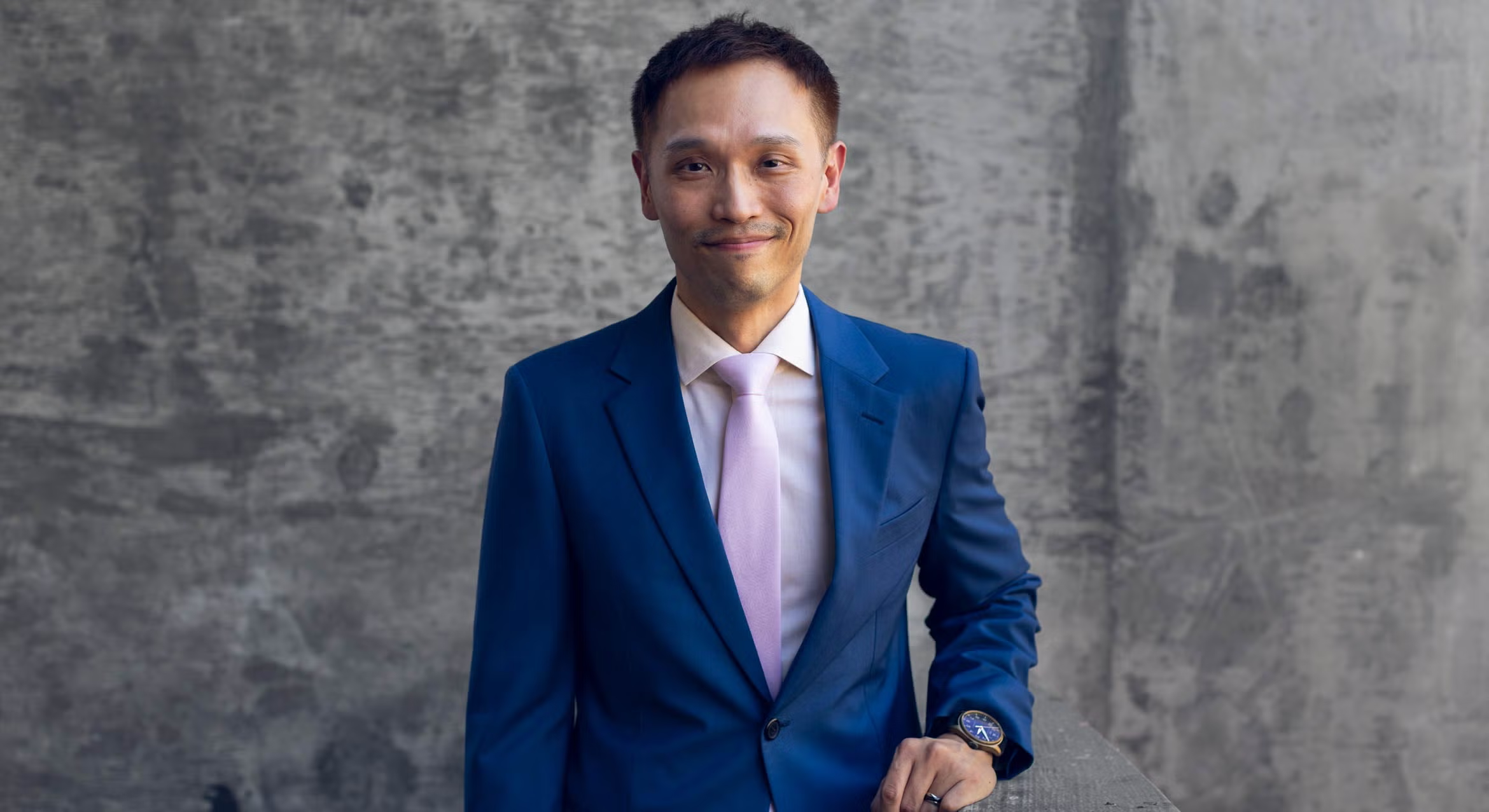 UCSB historian Howard Chiang smiling in a dark blue suit and light pink tie, standing outdoors against a textured concrete wall.