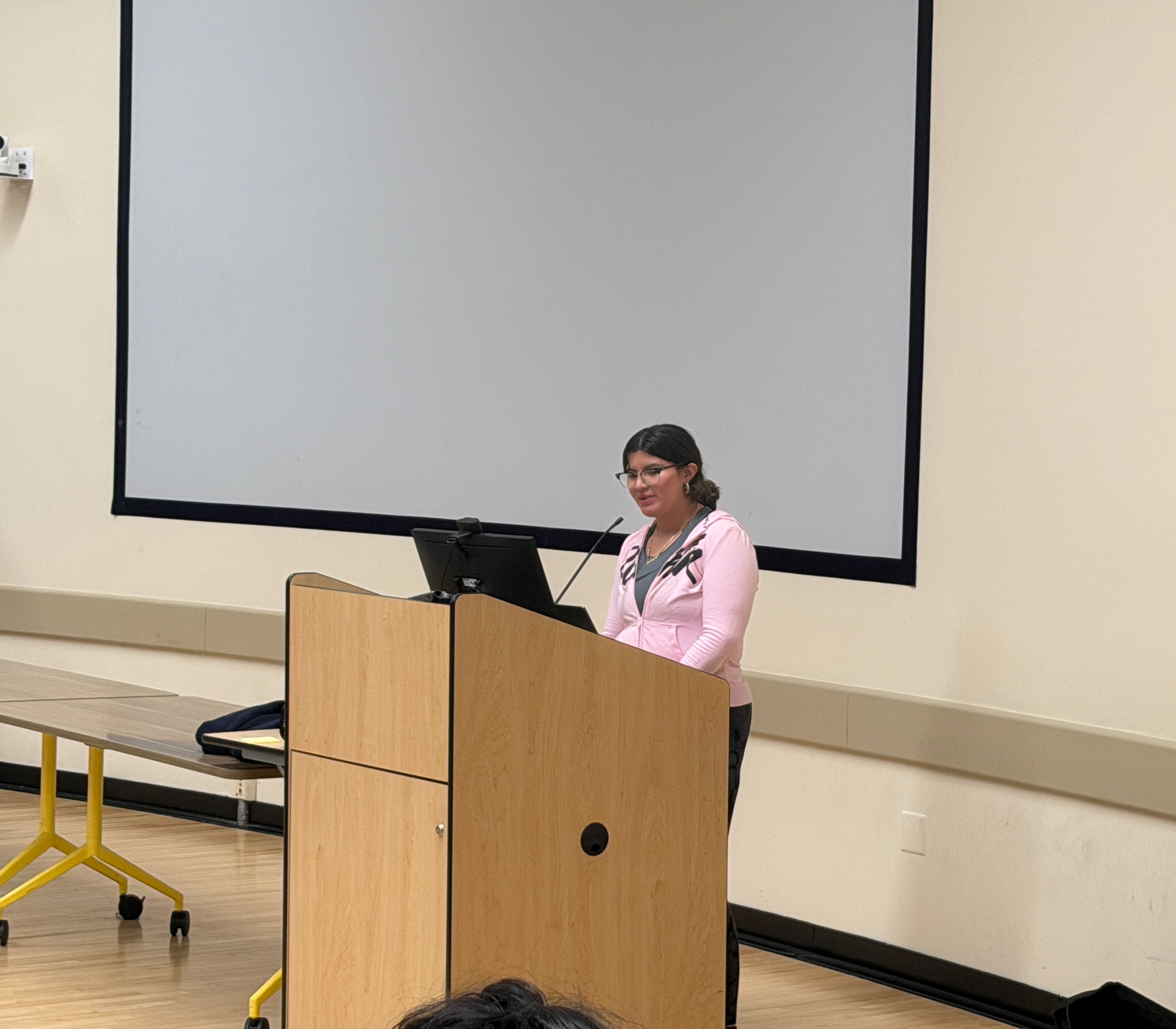 A young woman wearing glasses and a pink zip-up hoodie stands behind a light wooden podium, speaking into a microphone. A large, blank white projection screen hangs on the wall behind her.