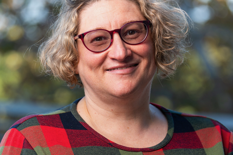 Professional headshot of Jenni Sorkin, smiling and wearing red-framed glasses and a multicolored plaid shirt.