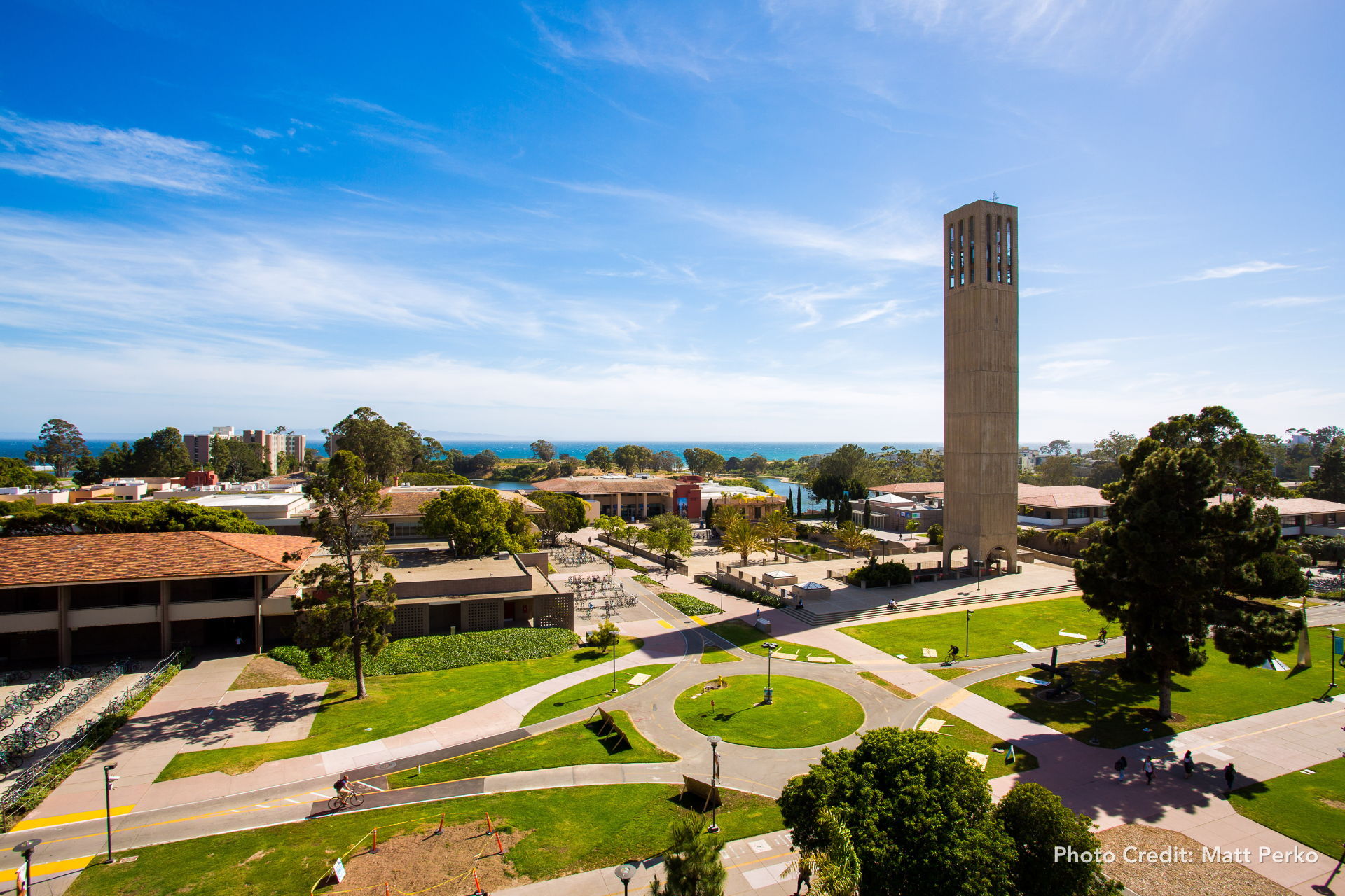 An aerial view of the University of California, Santa Barbara campus featuring the tall, rectangular Storke Tower rising above green lawns and surrounding academic buildings, with the Pacific Ocean and bright blue sky in the distance. Photo Credit: Matt Perko appears in lower right.