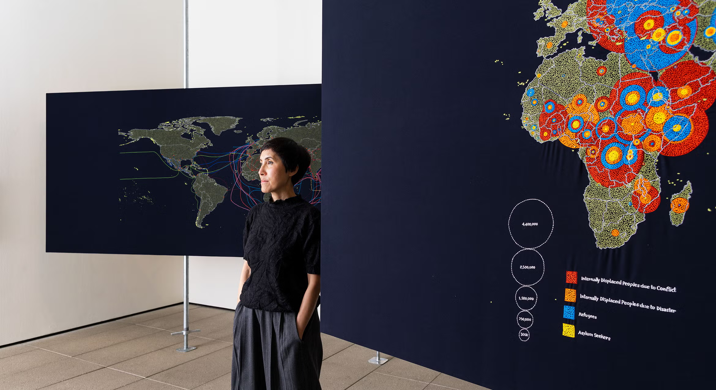 Artist Tiffany Chung stands in front of navy hand-embroidered maps in a museum gallery.