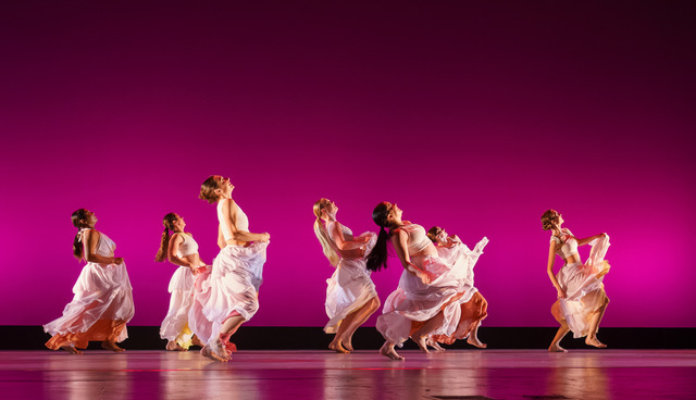 Seven dancers from the UCSB Dance Company perform "We Were Light" on stage against a vibrant magenta backdrop. They are wearing white dresses over orange skirts and are leaning backward in unison.