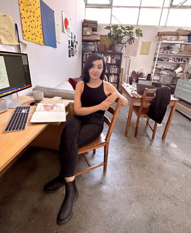 A full-body photograph shows Iman Djouini sitting sideways on a wooden chair. Her left arm rests on the back of the chair while her right arm is casually draped over her knee. She looks directly at the camera with a slight smile. She is in what appears to be an art studio, with a wooden desk and computer to her left. The studio also contains shelves filled with books and art supplies, and various artworks are visible on the walls and surfaces. 