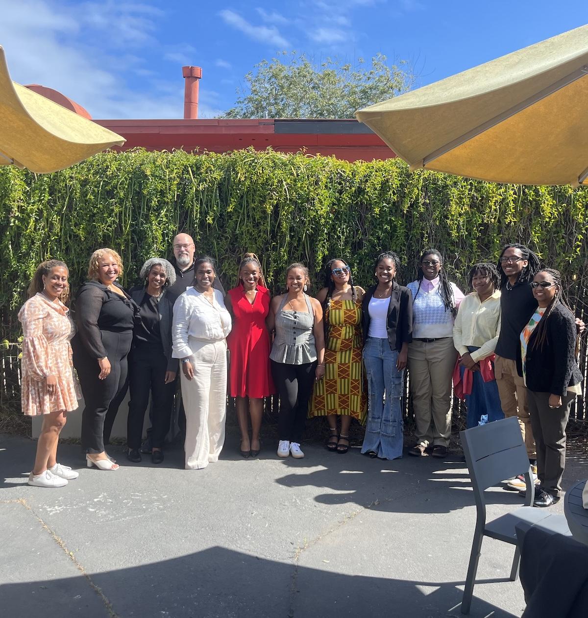 Group photo from a Booker T. Washington Scholars celebration in 2025. Fourteen individuals, including students and mentors, stand and smile for the camera outdoors in front of a green ivy wall, partially shaded by two large umbrellas.